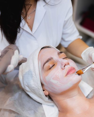 Beautician with a brush applies a white moisturizing mask to the face of a young girl client in a spa beauty salon